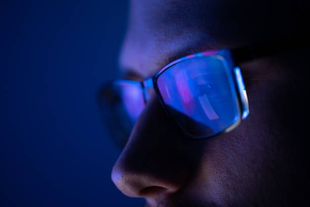 Close-up of a man wearing blue light blocking glasses with screen reflections visible on the lenses in a dark room.