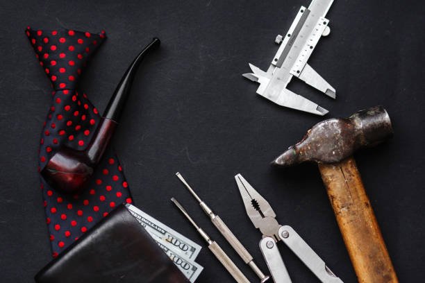 A flat-lay image on a black background featuring a red polka-dot tie with a smoking pipe, a black wallet with dollar bills, a caliper, a multitool with pliers and screwdrivers, and a hammer, arranged to symbolize a blend of style and utility for men.