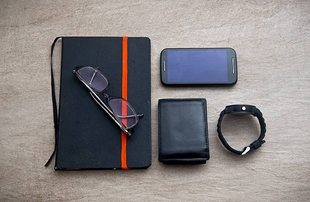 Flat lay of men's essentials including a notebook with glasses, a smartphone, black leather wallet, and a smartwatch on a stone surface.