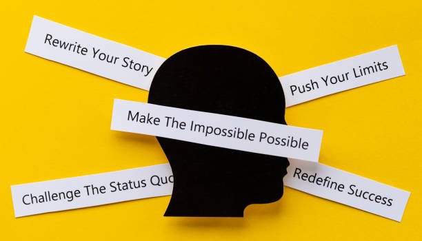 A black silhouette of a human head in profile on a bright yellow background, with five white paper strips radiating outward. Each strip contains a motivational phrase: "Rewrite Your Story," "Push Your Limits," "Redefine Success," "Challenge the Status Quo," and "Make the Impossible Possible."