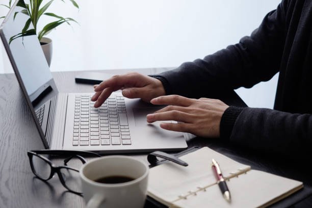 A man working on a laptop at a wooden desk, surrounded by a notebook, pen, coffee mug, and glasses, symbolizing a focused side hustle environment.
