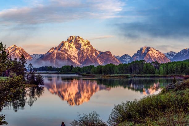 A serene lake reflecting snow-capped mountains at sunrise, with a person standing by the water in a national parks landscape.
