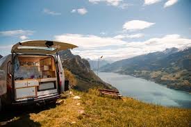 A converted van with an open rear door, parked on a scenic mountain overlook with a lake and peaks in the background, showcasing a van life setup.