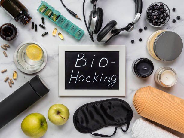 A flat lay image featuring a "Bio-Hacking" sign surrounded by supplements, a water glass with lemon, headphones, a yoga mat, sleep mask, apples, a water bottle, candles, and a pill organizer, symbolizing cognitive biohacking techniques.