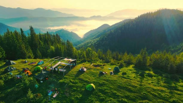 Aerial view of a mountain campsite at sunrise, with tents scattered on a grassy hill overlooking a vast, forested valley indicating a national park.