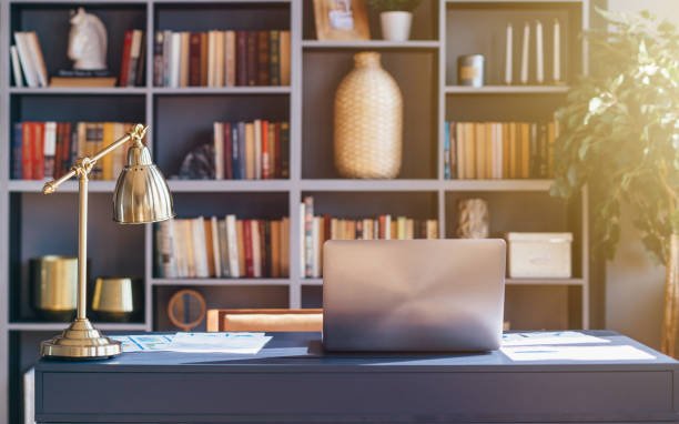 A stylish home office setup with a dark desk holding a laptop, a gold desk lamp, and papers, set against a bookshelf filled with books, decorative items, and a plant, illuminated by natural sunlight.