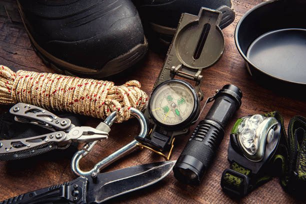 A close-up of essential outdoor gear for a camping checklist laid out on a wooden table, including hiking boots, a compass, flashlight, headlamp, rope, and a multi-tool.