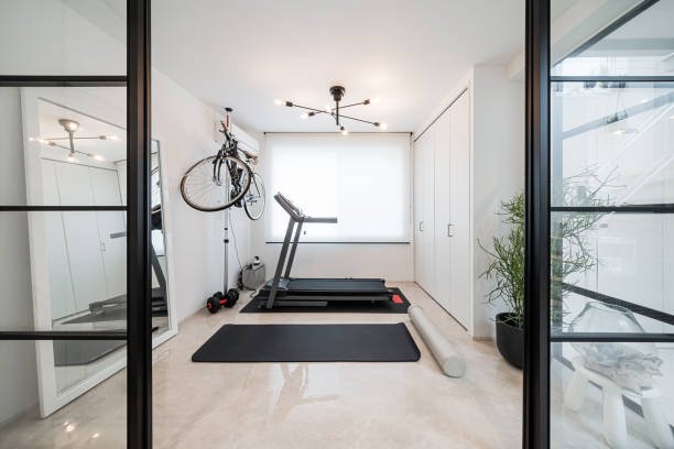 A compact and modern home gym setup in a bright room featuring a treadmill, yoga mat, dumbbells, and a wall-mounted bicycle.