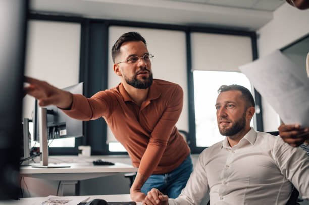 Two men collaborating in a modern office, one pointing at a computer screen while the other holds documents, symbolizing mentorship and professional growth for men over 30 in 2025.