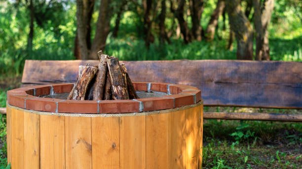 A wooden outdoor fire pit with a brick rim and burning logs, set in a grassy area with trees and a wooden bench in the background.