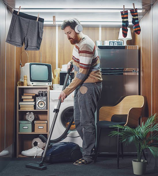 A man vacuuming a compact tiny home with a washing machine, fridge, and minimal furniture, showcasing masculine minimalist living.
