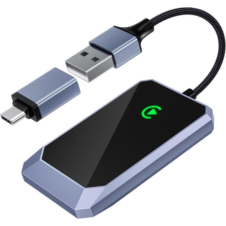 A sleek grey and black wireless CarPlay adapter with a glowing green connection light, shown with its attached USB cable and a USB-C adapter.