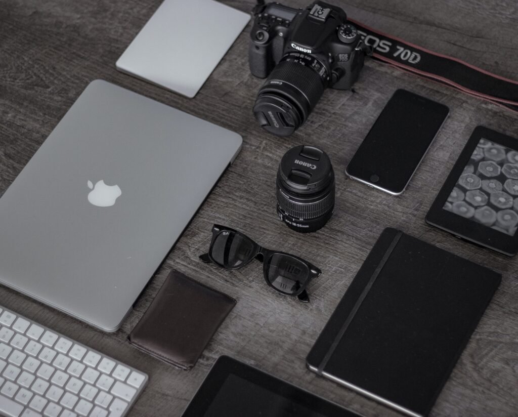 A flat lay of luxury tech gadgets, including a MacBook, Canon camera, and smartphones, neatly arranged on a wooden desk to represent a functional modern toolkit.