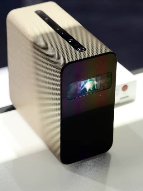 A stylish, compact mini projector with a gold perforated casing and a sleek black face, shown sitting on a white surface.
