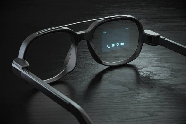 A pair of modern black smart glasses on a dark table, with a glowing blue heads-up display showing call, message, and camera icons on the lens.