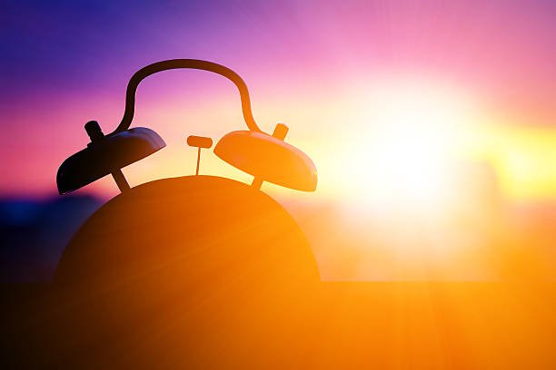 A classic twin-bell alarm clock silhouetted against a beautiful, vibrant sunrise, representing a natural awakening.