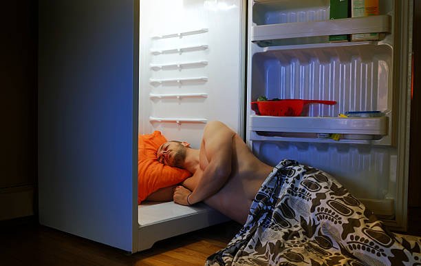 Man sleeping uncomfortably in an open refrigerator with an orange pillow, looking for extreme cooling on a hot night. Highlights the desperate need for a cooling pillow sleep solution.