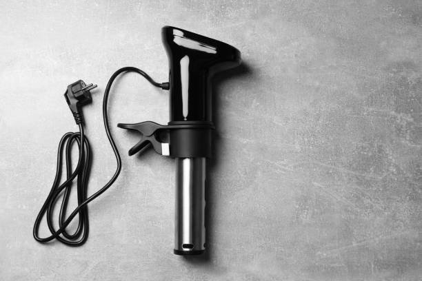 Black and white image of a sleek Anova-style sous vide circulator with a power cord, lying on a grey concrete surface.