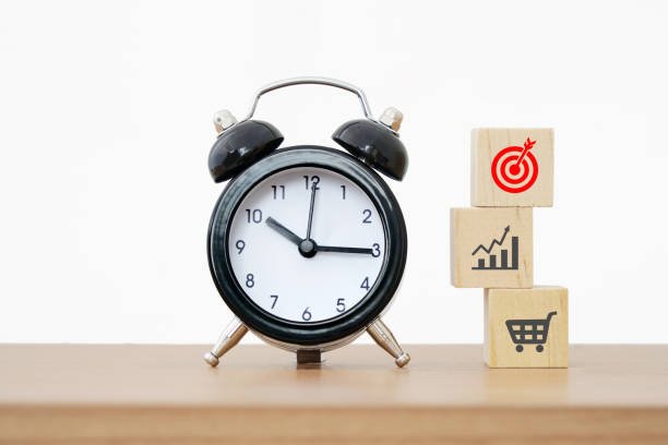 Analog alarm clock next to wooden blocks showing productivity icons: target goal, growth chart, and shopping cart. Focus on analog tools for productivity.